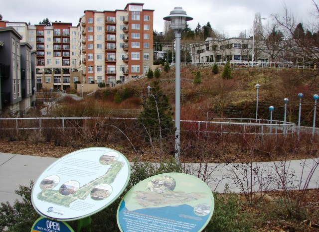Looking east across the Thornton Creek Water Quality Channel, we see the pathways and viewing areas around the channel cut diagonally through the block. The buildings on the perimeter are: left: Thornton Place Apartments; center: Aljoya retirement community; right: Group Health building on the corner of 5th Ave NE and NE 100th Street.