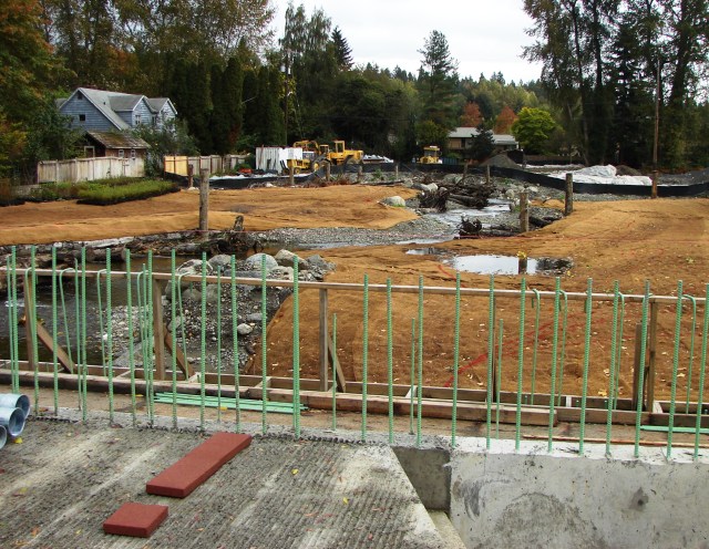 Looking eastward from 35th Ave NE, in the foreground we see the road repair work with sidewalk and rails, which will give this section a bridge-like appearance.  A flood plain is being created as the South Branch of Thorton Creek flows toward Meadowbrook Pond.  The house at left faces 36th Ave NE.
